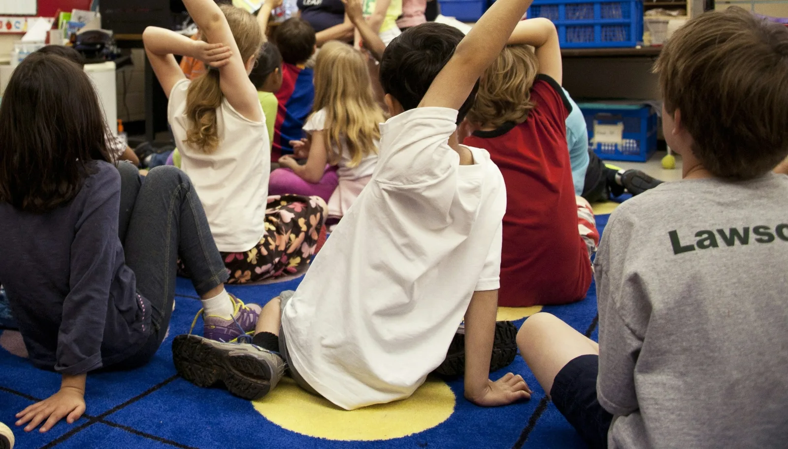Children sitting on a classroom floor with their hands raised, seen from behind
